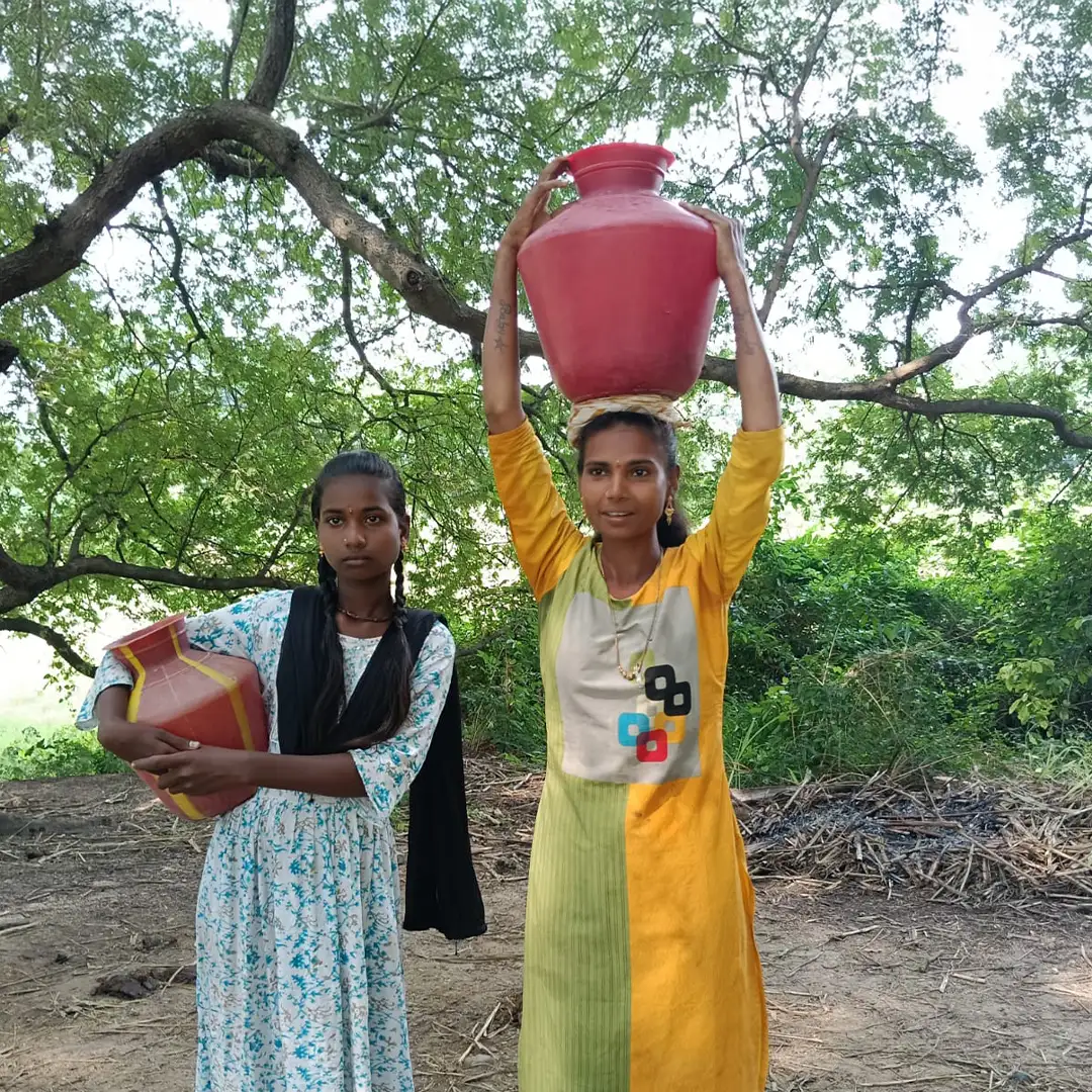 Two girls standing in the shade of a tree. One holds a clay water jar, the other balances it atop her head.